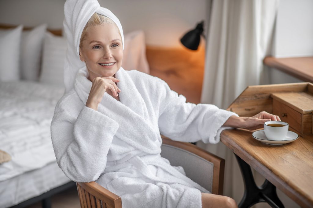 A mature woman in a white robe enjoying a cup of tea after rejuvenating skincare in her luxurious home spa with SQOOM